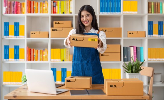 Woman handing out a GLS parcel. She stands at a desk in front of shelves field with colourful books. Woman handing out a GLS parcel. She stands at a desk in front of shelves field with colourful books.